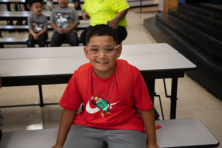 A boy in glasses and a red shirt smiles as he sits at a cafeteria table