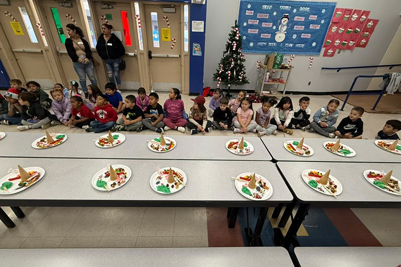 Students sit on the cafeteria floor, with tables filled with Christmas tree decorations