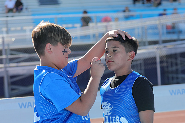 A boy in a blue shirt paints another boys face with a black marker