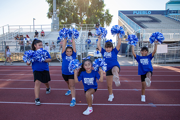 Girls in blue shirts holding blue and white pom poms perform a cheer on the sidelines