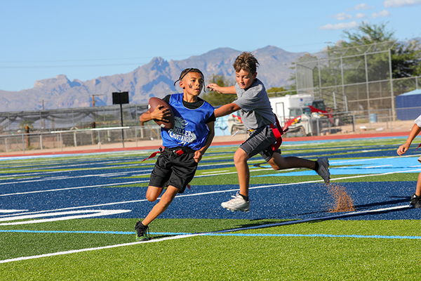 A boy in a blue jersey holding a football runs from a boy in a gray shirt